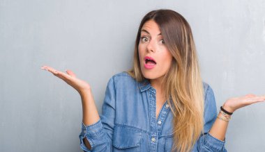 Grunge gri duvar denim kıyafet clueless ve şaşkın bir ifadeyle kollar ve eller kaldırdı giyen genç yetişkin kadın. Şüphe kavramı.