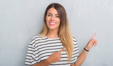 Grunge gri duvar gülümseyen ve iki elleri ve parmakları ile tarafına işaret kamera bakarak lacivert t-shirt giyen genç yetişkin kadın.