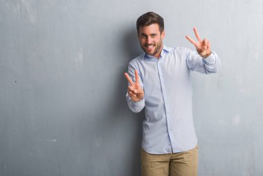 Yakışıklı genç iş adamı zafer işareti yapıyor parmak gösterilen kamera seyir gülümseyen gri grunge duvar giyen şık gömlek üzerinde. İki numaralı.