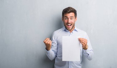 Yakışıklı genç gri grunge duvar holding boş kağıt sayfası üzerinden sözleşme gurur çığlık ve kutluyor zafer ve başarı çok heyecanlı, duygu tezahürat