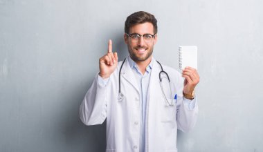 Yakışıklı genç doktor adam gri grunge boş defter sürpriz bir fikir ile tutarak duvar veya gülen yüz, bir numara ile işaret parmağı soru