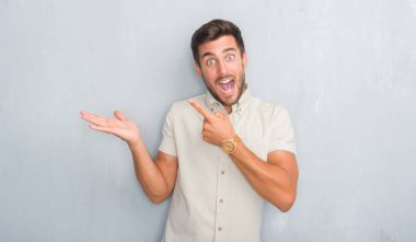 Yakışıklı genç gri grunge üzerinde duvar giyen Yaz gömlek şaşırdım ve el ile sunulması ve parmak ile işaret ederken kameraya gülümseyen.