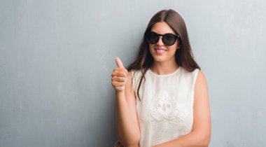 Grunge genç esmer kadın ok işareti, başparmak ile parmak, mükemmel işareti yapıyor kocaman bir gülümseme ile duvar giyen moda güneş gözlüğü mutlu gri