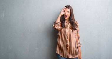 Grunge genç esmer kadın gri duvar ok jest sürpriz yüz, göz parmaklarının arasından seyir şok yapıyor. İmansız ifade.