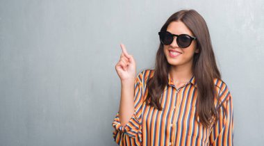 Grunge gri genç esmer kadın giyiyor büyük güneş gözlüğü bir fikirle sürpriz duvar veya gülen yüz, bir numara ile işaret parmağı soru