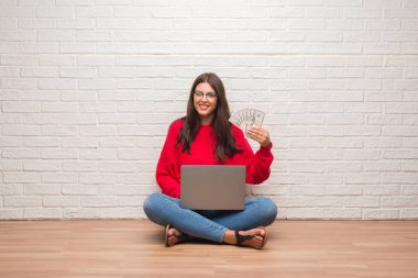 Ayakta ve diş gösterilen kendine güvenen bir gülümsemeyle gülümseyerek mutlu bir yüz ile holding dolar ödemek beyaz tuğla duvarın üzerinden katta oturan genç esmer kadın