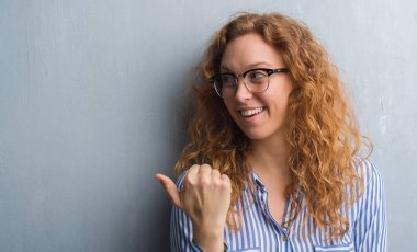 Genç Kızıl saçlı kadın işaret ve başparmak kadar mutlu yüz gülümseyen tarafı ile gösterilen gri grunge duvarın üzerinden