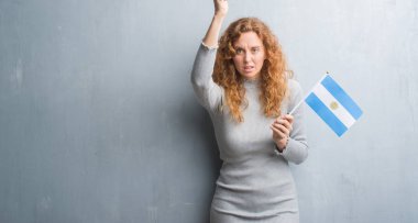 Gri grunge duvar rahatsız Arjantin bayrağı ve öfke, çılgın ve yükseltilmiş el, öfke kavramı ile diye bağıran ile sinirli bağırarak tutan genç Kızıl saçlı kadın