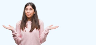 Young beautiful hispanic woman wearing a sweater clueless and confused expression with arms and hands raised. Doubt concept. clipart