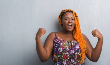 Genç Afro-Amerikan kadın turuncu giymiş gri grunge duvarın üzerinden kaldırdı kollarını açarak şaşırmış ve hayret için başarı kutluyor örgü ve gözleri açık. Kazanan kavramı.