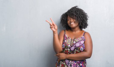 Gri grunge duvar renkli giyen genç Afro-Amerikan kadın elbise mutlu yüz zafer işareti yaparken kameraya göz kırpıyor ile gülümseyen. İki numaralı.