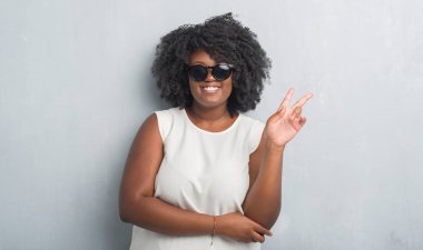 Genç Afro-Amerikan artı boyutu kadın moda mutlu yüz zafer işareti yaparken kameraya göz kırpıyor ile gülümseyen güneş gözlüğü takıyor gri grunge duvarın üzerinden. İki numaralı.