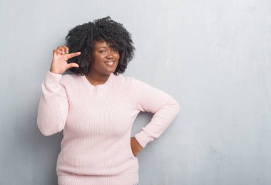 Genç Afro-Amerikan artı boyutu kadın kış kazak gülümseyen ve süre seyir ve kamera boyutu işaret parmaklarıyla yapıyor el ile kendinden emin eller gri grunge duvarın üzerinden. Ölçü kavramı.