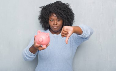 Kızgın yüz, eksi işareti aşağı antipati başparmak ile gösterilen, ret kavramı ile piggy banka holding gri grunge duvarın üzerinden genç Afro-Amerikan kadın
