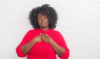 Genç Afro-Amerikan artı boyutu kadın kapalı gözler ile göğüs ellerinde ve yüzünde minnettar jest ile gülümseyen beyaz tuğla duvarın üzerinden. Sağlık kavramı.