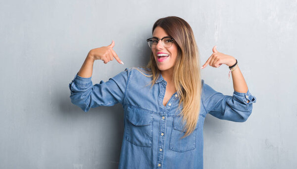 Young adult woman over grunge grey wall wearing glasses looking confident with smile on face, pointing oneself with fingers proud and happy.