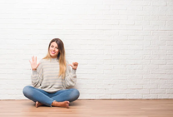 年轻的成年女子坐在地板上的白色砖墙在家里显示和指向六的手指 而微笑着自信和快乐 — 图库照片