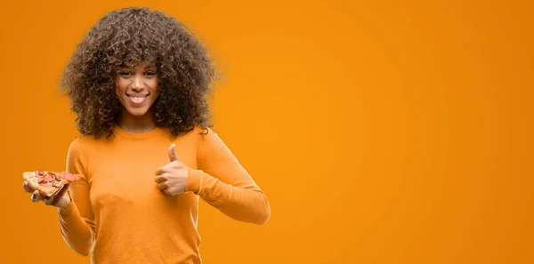 African american woman with a pizza slice happy with big smile doing ok sign, thumb up with fingers, excellent sign