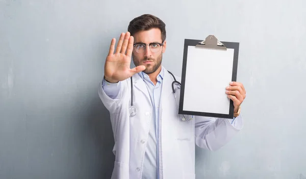 英俊的年轻医生人在灰色垃圾墙持有剪贴板与张开手做停止标志认真和自信的表达 防御姿态 — 图库照片
