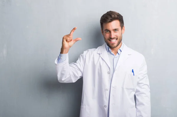 Handsome young professional man over grey grunge wall wearing white coat smiling and confident gesturing with hand doing size sign with fingers while looking and the camera. Measure concept.