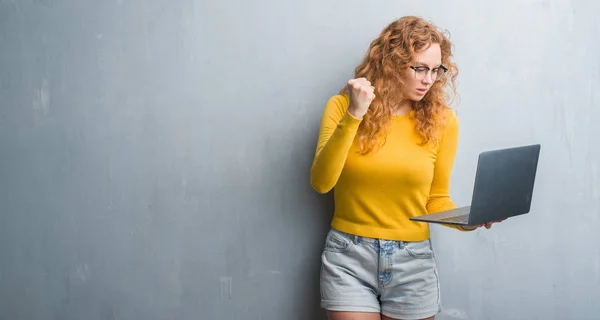 灰色垃圾墙上的年轻红头发的女人拿着电脑笔记本电脑的恼怒和沮丧的呼喊与愤怒 疯狂和叫喊与举手 愤怒的概念 — 图库照片