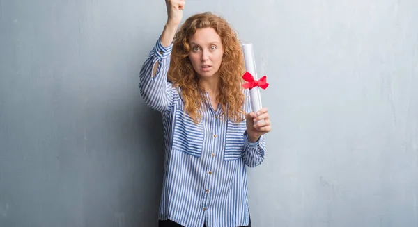 Young redhead business woman over grey grunge wall holding diploma annoyed and frustrated shouting with anger, crazy and yelling with raised hand, anger concept