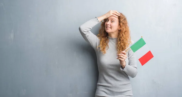 Young redhead woman over grey grunge wall holding flag of Italy stressed with hand on head, shocked with shame and surprise face, angry and frustrated. Fear and upset for mistake.