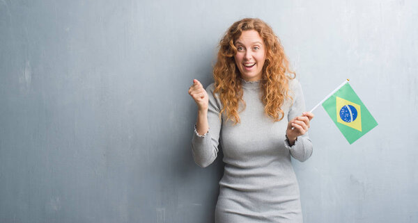 Молодая рыжая женщина над серой гранж-стеной с флагом Бразилии, гордо кричащая и празднующая победу и успех, очень взволнована, аплодирует эмоциям

