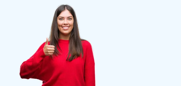 若い美しいヒスパニック系ハッピー サムズアップ手でジェスチャーをしている赤いセーターを着ています 成功を示すと カメラを見て式を承認します — ストック写真