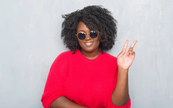 Young african american plus size woman over grey grunge wall wearing retro sunglasses smiling with happy face winking at the camera doing victory sign. Number two.