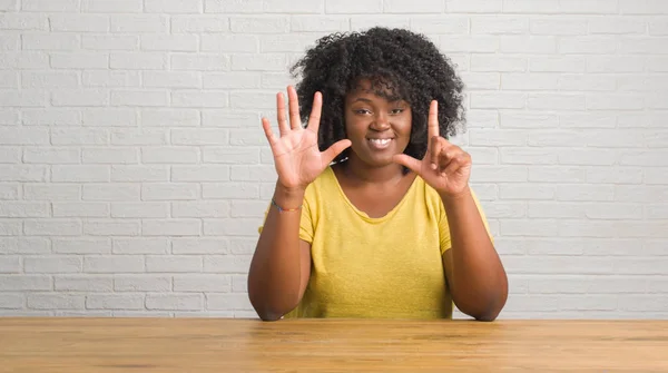 Young african american woman sitting on the table at home showing and pointing up with fingers number seven while smiling confident and happy.