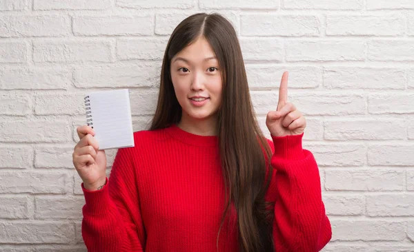 Joven Mujer China Sobre Pared Ladrillo Mostrando Cuaderno Blanco Sorprendido —  Fotos de Stock