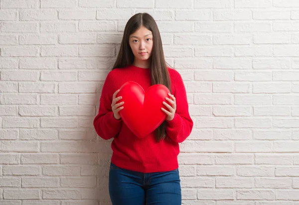 Young Chinese woman in love over brick wall holding red heart with a confident expression on smart face thinking serious