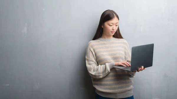 年轻的中国妇女在灰色墙壁使用计算机膝上型电脑与自信的表情在聪明的面孔认真思考 — 图库照片