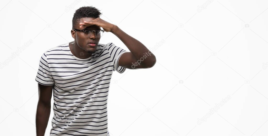 Young african american man wearing glasses and navy t-shirt very happy and smiling looking far away with hand over head. Searching concept.