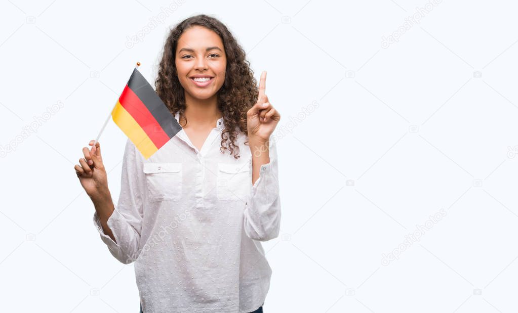 Young hispanic woman holding flag of Germany surprised with an idea or question pointing finger with happy face, number one