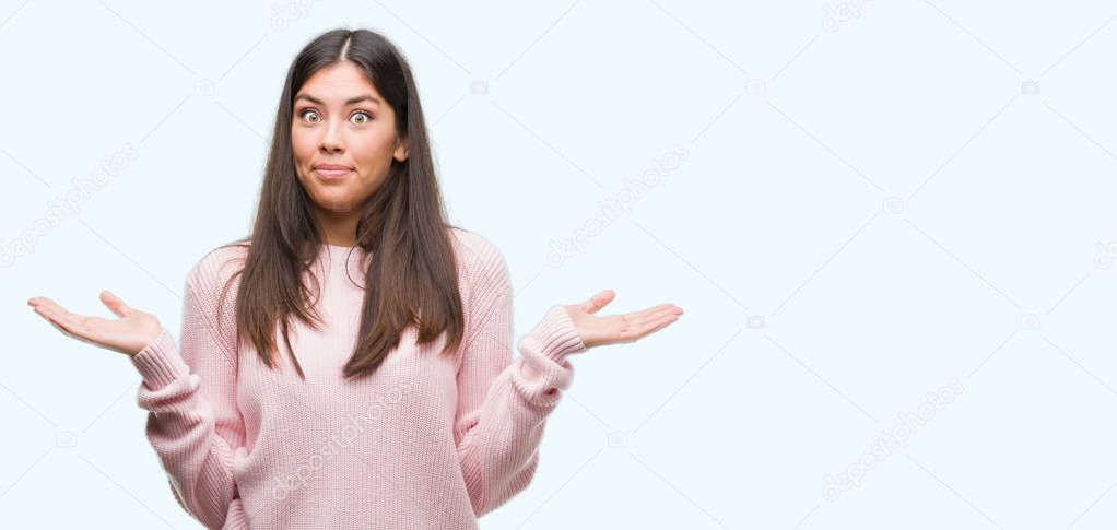 Young beautiful hispanic woman wearing a sweater clueless and confused expression with arms and hands raised. Doubt concept.