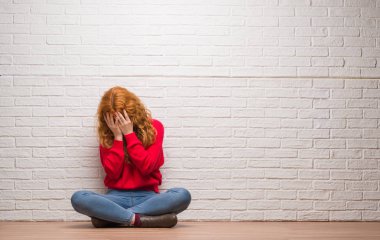 Yüzünü elleriyle ağlayarak süre kapsayan üzgün ifade ile tuğla duvarın üzerinden oturan genç Kızıl saçlı kadın. Depresyon kavramı.