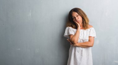 Gri grunge duvar düşünme seyir duran orta yaş İspanyol kadın yorgun ve sıkılmış çapraz silah depresyon sorunları ile.