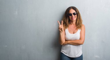 Orta yaş İspanyol kadın gri duvar mutlu yüz zafer işareti yaparken kameraya göz kırpıyor ile gülümseyen güneş gözlüğü takıyor. İki numaralı.