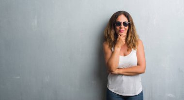 Orta yaş İspanyol kadın gri duvar emin çapraz kollar ve çeneme kaldırdı el ile gülümseme ile kameraya bakarak güneş gözlüğü takıyor. Pozitif düşünce.