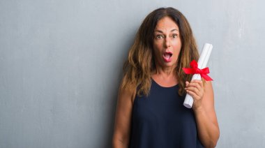 Diploma korkmuş şok bir sürpriz yüz, korkuyor ve heyecanlı korku ifadesi ile tutarak gri grunge duvarın üzerinden duran orta yaş İspanyol kadın