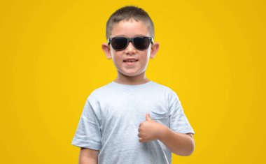 Koyu saçlı küçük çocuk giyen güneş gözlüğü sürpriz ile işaret parmağı kendisine yüz