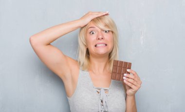 Baş, el ile vurguladı çikolata yeme gri grunge duvarın üzerinden Kafkas yetişkin kadın şok utanç ve sürpriz suratla, öfkeli ve sinirli. Korku ve hata için üzgün.