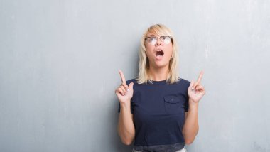 Yetişkin kafkas kadın gözlük takan grunge gri duvarın üzerinden hayran ve yukarıya bakıyor ve parmakları ile işaret ve silah kaldırdı şaşırttı.