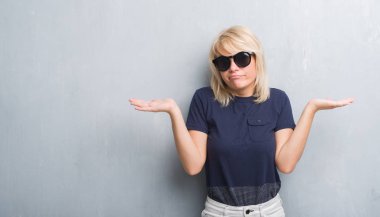 Yetişkin beyaz kadın güneş gözlüğü clueless ve şaşkın bir ifadeyle kollar ve eller kaldırdı giyen grunge gri duvar yüzünden. Şüphe kavramı.