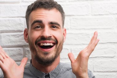 Kocaman bir gülümseme ile çığlık zaferini kutluyor beyaz tuğla duvar çok mutlu ve heyecanlı, kazanan ifade üzerinde duran ve ellerini kaldırdı genç yetişkin adam