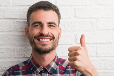 Genç yetişkin adam beyaz tuğla duvardan mutlu büyük gülümseme ok işareti, başparmak ile parmak, mükemmel işareti yapıyor ayakta