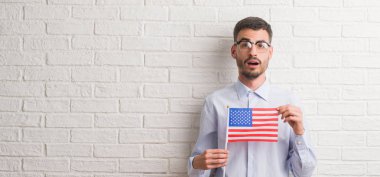 Genç yetişkin adam Amerika bayrağı korkmuş şok bir sürpriz yüz, korkuyor ve heyecanlı korku ifadesi ile tutarak tuğla duvarın üzerinden