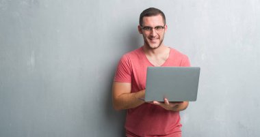 Genç beyaz adam ayakta ve diş gösterilen kendine güvenen bir gülümsemeyle gülümseyerek mutlu bir yüz ile bilgisayar dizüstü kullanarak gri grunge duvar üzerinde